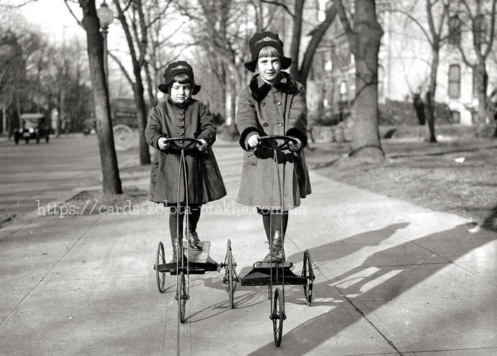 Two girls in coats and caps riding four-wheeled pedal cars on a tree-lined sidewalk