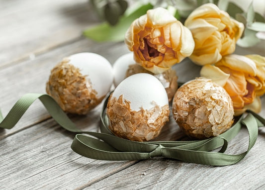 White eggs half-covered in gold foil flakes beside a green ribbon and apricot tulips