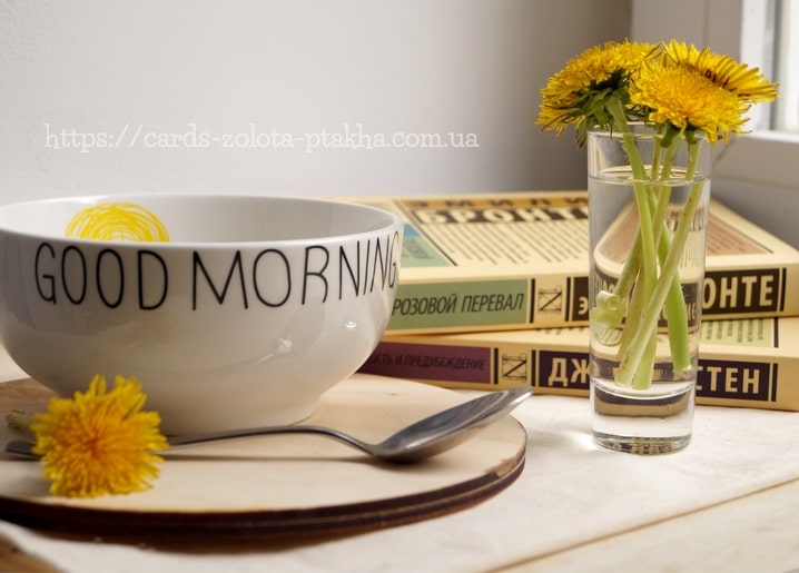 Weiße „Guten Morgen“-Schale mit Löffel und Löwenzahn im Glas vor Büchern