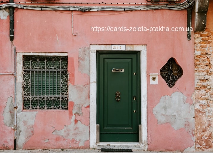 Verwitterte rosa Wand mit dunkelgrüner Tür, Hausnummer 2114 und schmiedeeisernem Fenstergitter.