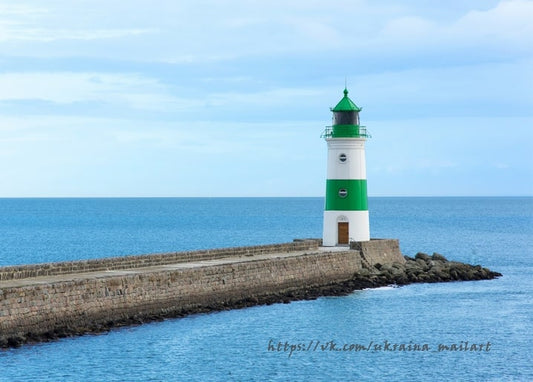 Grün-weißer, gestreifter Leuchtturm am Ende einer Steinmole vor ruhigem blauem Meer und blassem Himmel