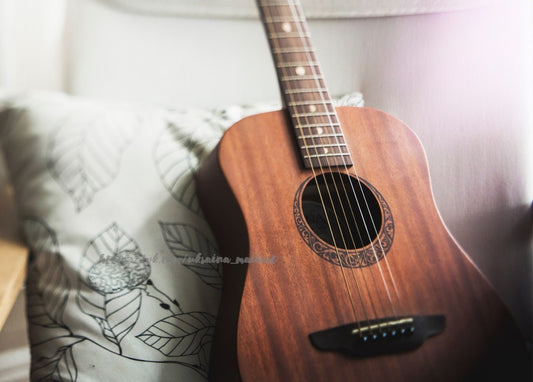 Akustikgitarre mit Mahagoni-Korpus lehnt im weichen Tageslicht an einem Kissen mit Blattmuster
