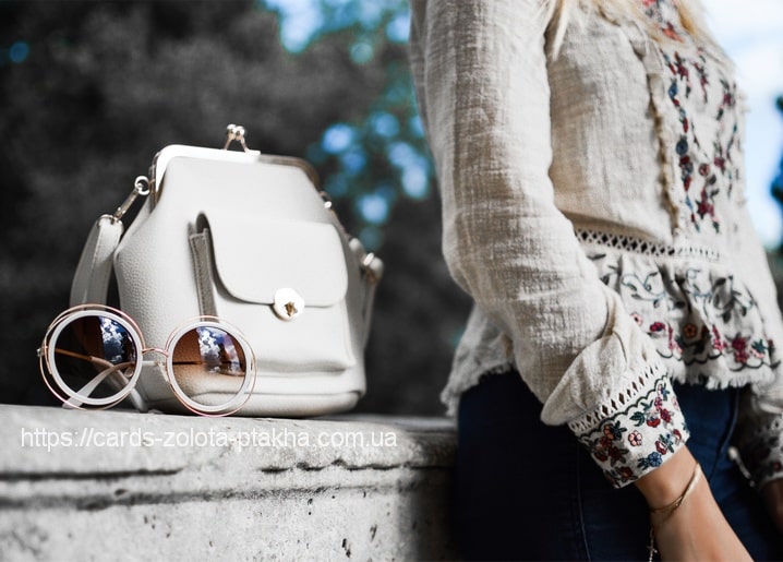 Weiße Handtasche und runde Sonnenbrille mit roségoldenem Rahmen auf einer Steinmauer