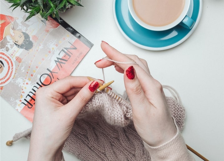 Hands knitting a beige sweater beside coffee and an open magazine