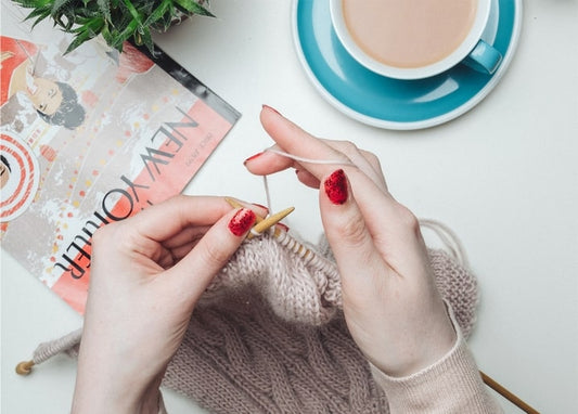 Hands knitting a beige sweater beside coffee and an open magazine