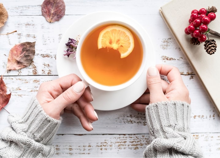 Hands holding a cup of lemon tea over a table with dried leaves and berries