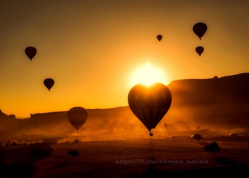 Herzförmiger Heißluftballon leuchtet im Sonnenaufgang, weitere Ballons über nebligen Hügeln
