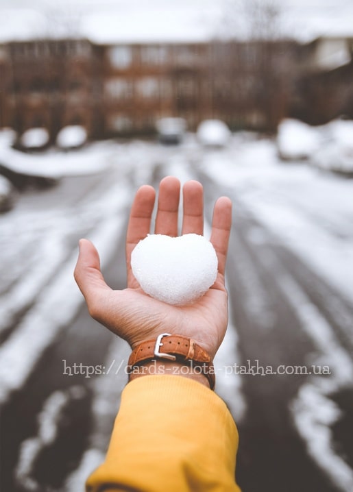 Hand hält einen herzförmigen Schneeball vor einer verschneiten Straße