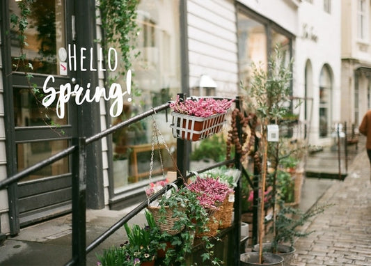 Shop storefront with hanging flower baskets, potted olive trees, and a “Hello Spring!” sign