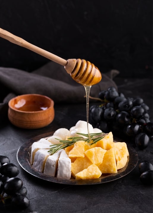 Honey drizzling from a dipper over camembert and cheese cubes with black grapes