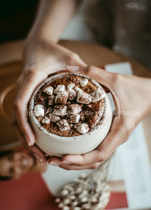 Zwei Hände halten eine Tasse heiße Schokolade mit Marshmallows und Kakaopulver