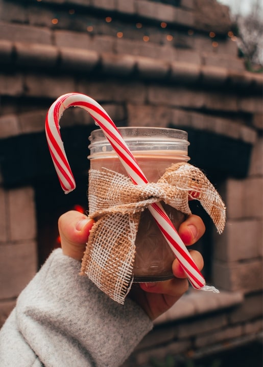 Hand hält ein mit Sackleinen umwickeltes Glas heißem Kakao mit Zuckerstange vor einem Steinkamin