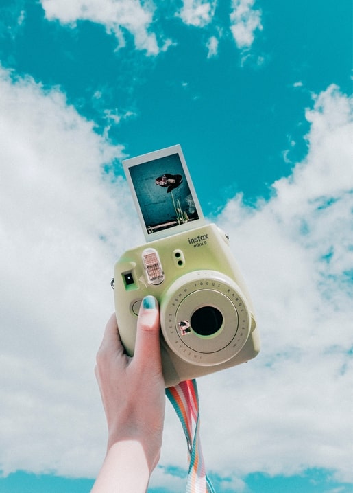 Hand hält eine Instax-Sofortbildkamera vor blauem Sommerhimmel mit Wolken