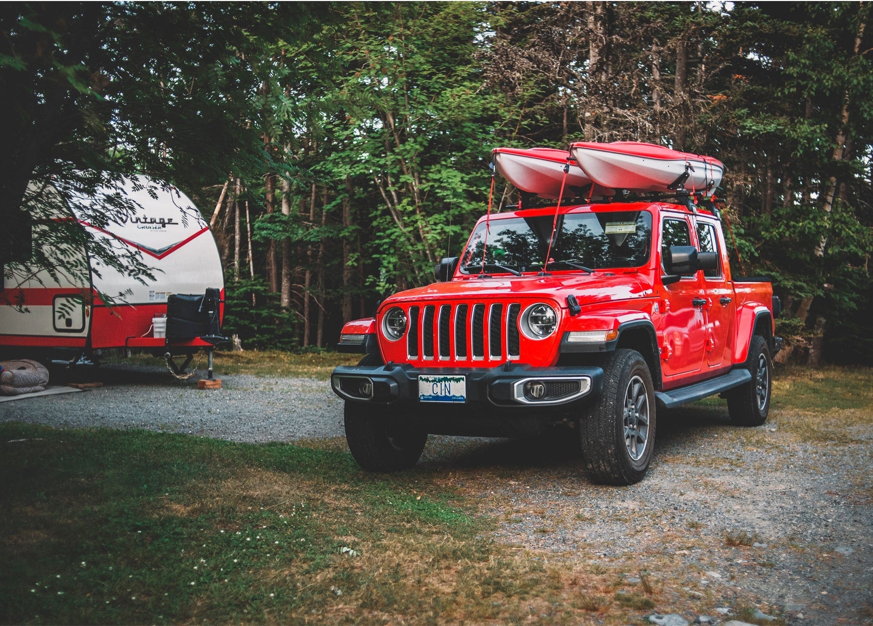 Roter Jeep Gladiator mit zwei Kajaks auf dem Dach neben einem Vintage-Wohnwagen im Wald