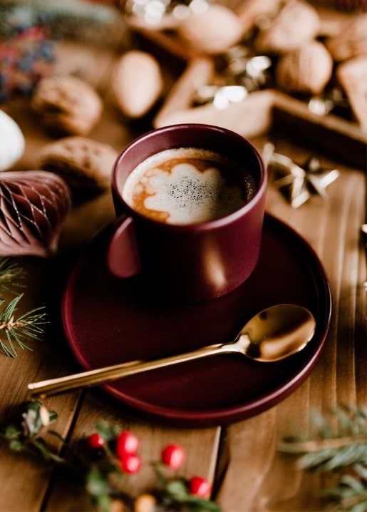 Latte with foam art on a burgundy saucer beside a gold spoon and pine sprig