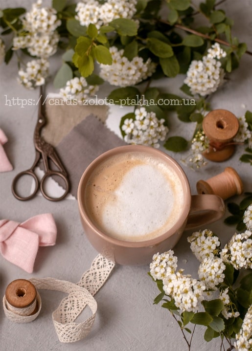 Rosa Latte mit Milchschaum neben Vintage-Schere, Garnrollen aus Holz und weißen Frühlingsblüten.
