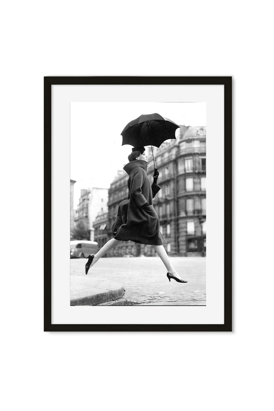 Black-and-white photo of a woman leaping with an open umbrella across a Paris street, framed poster