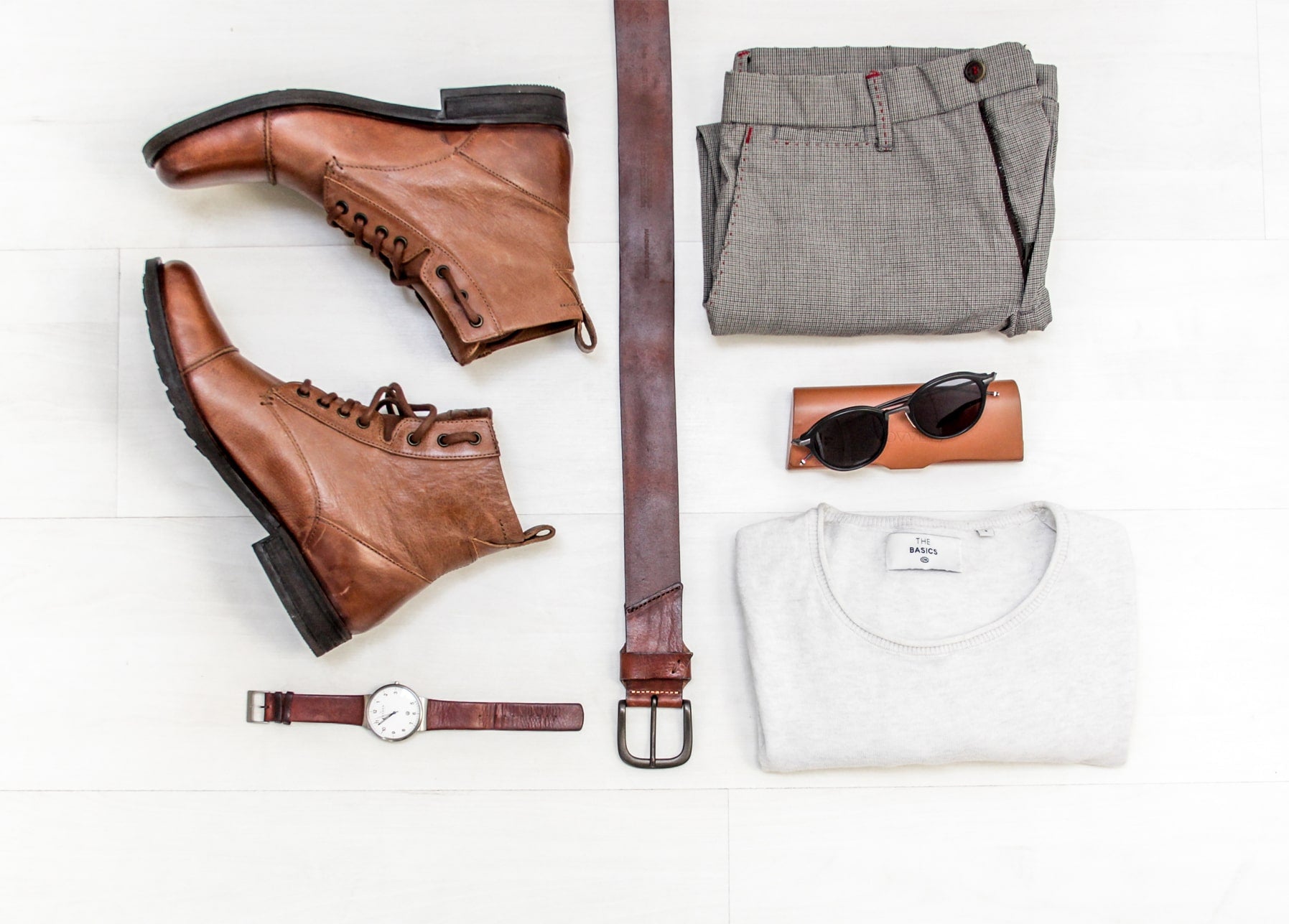 Flatlay aus Lederstiefeln, Gürtel, Uhr, Sonnenbrille und gefaltetem grauem Pullover