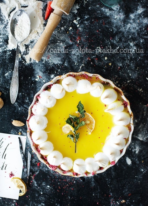 Lemon meringue tart with lemon slices, photographed flat-lay among flour and a rolling pin