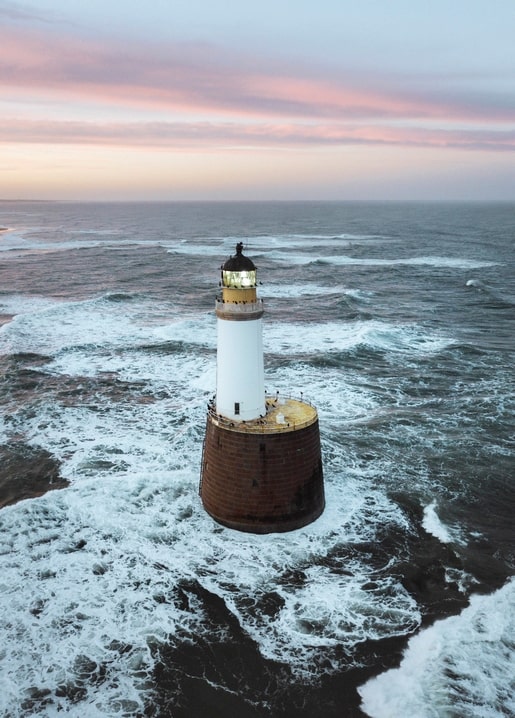 Lighthouse surrounded by crashing waves under a pink sunset sky