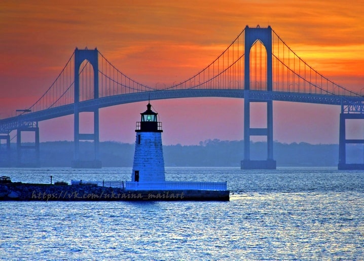 Kleiner Steinleuchtturm auf einer Mole, dahinter eine große Hängebrücke bei Sonnenuntergang