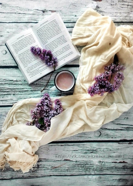 Гілочки бузку, розгорнута книга та чашка чаю на дерев'яному столі, огорнуті кремовим шарфом