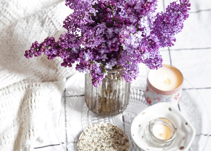 Purple lilac in a glass jar beside lit candles and lace on a table