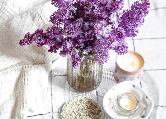 Purple lilac in a glass jar beside lit candles and lace on a table