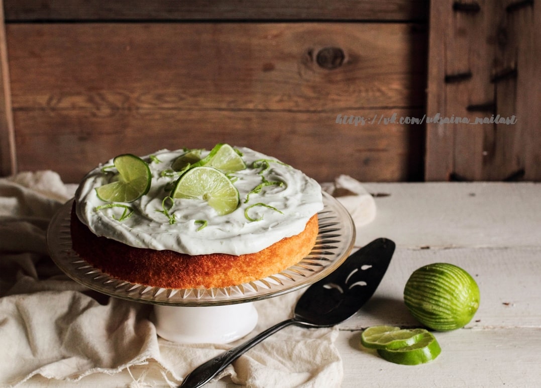 Frosted lime cream cake on a glass stand with lime slices, a whole lime and wedges nearby