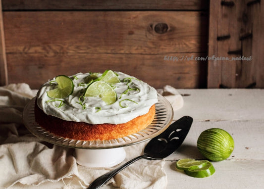 Frosted lime cream cake on a glass stand with lime slices, a whole lime and wedges nearby