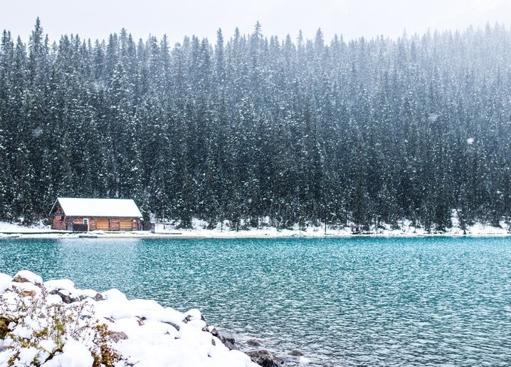 Blockhütte am Ufer eines türkisfarbenen Sees mit Schneefall im Kiefernwald