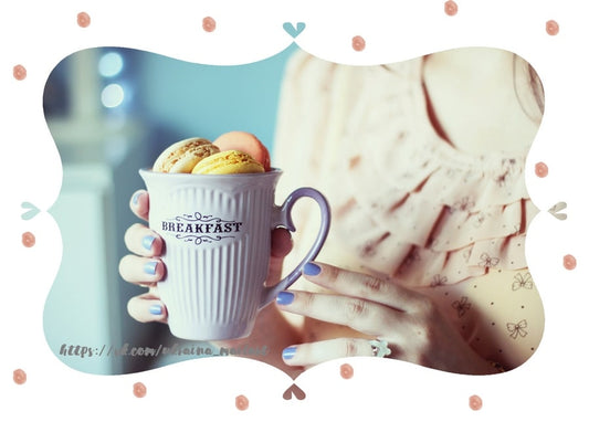 Hand mit lila Nagellack hält eine weiße „Breakfast“-Tasse voller bunter Macarons