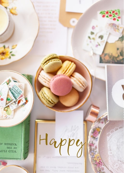 Flat lay of pastel macarons, postage stamps, and floral stickers beside a gold ‘Happy’ script