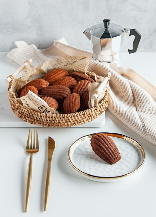Basket of golden madeleines beside a stovetop moka pot, gold cutlery, and a linen napkin on a white table
