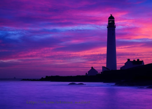 Weißer Leuchtturm als Silhouette vor magenta-violettem Sonnenuntergang über einer Felsenküste
