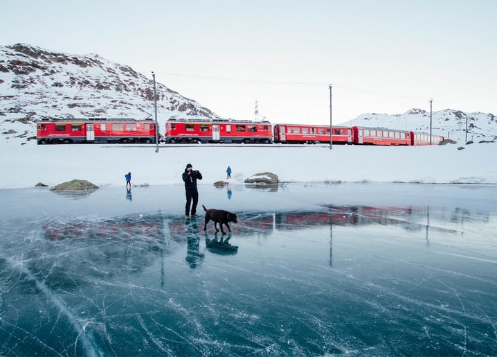 Man walking a brown dog across a cracked frozen lake with a red mountain train behind them