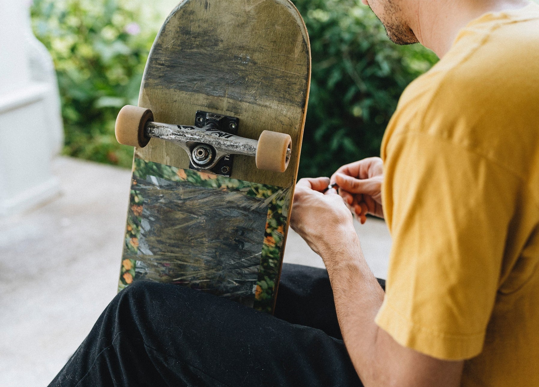 Man in a yellow t-shirt adjusting the trucks of a painted skateboard outdoors