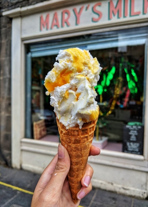 Hand hält ein Waffelhörnchen mit Vanille-Mango-Softeis vor dem Schaufenster einer Eisdiele.