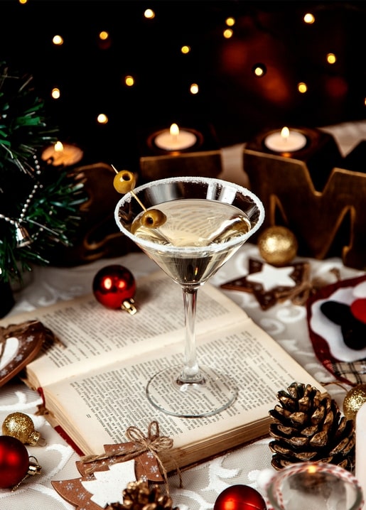 Martini cocktail with olives on an open book, surrounded by candles and gold pinecones.