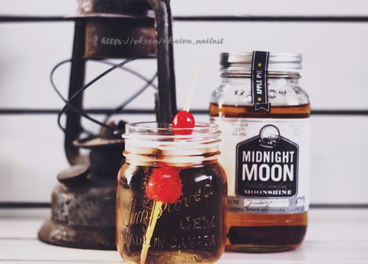 Cocktail im Einmachglas mit Kirschen neben einer Flasche Apple-Pie-Moonshine und einer alten Öllampe