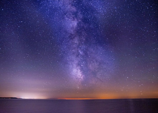 Milchstraße und Sterne über dunklem Ozean mit Lichtschein am Horizont