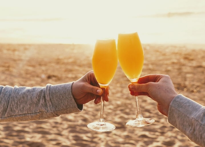Zwei Hände stoßen mit Mimosa-Gläsern am Strand bei Sonnenuntergang an
