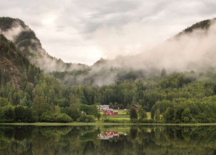 Nebliges Bergtal mit bewaldeten Hängen und einem roten Bauernhaus am See