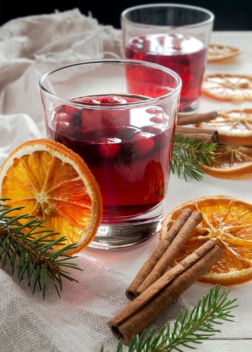 Zwei Gläser Glühwein mit Preiselbeeren neben Zimtstangen und getrockneten Orangenscheiben