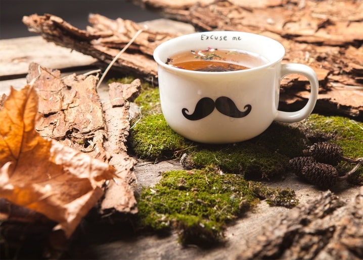 Mustache-print mug of tea on moss with autumn leaves and pinecones