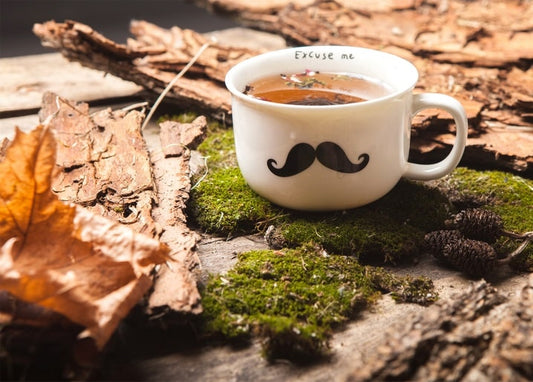 Mustache-print mug of tea on moss with autumn leaves and pinecones