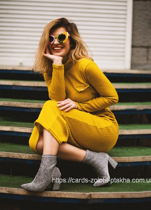 Lachende Frau im senffarbenen Strickkleid mit bunter Sonnenbrille auf Stadionstufen