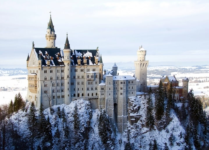 Neuschwanstein Castle covered in snow above a pine forest, mountains in the background