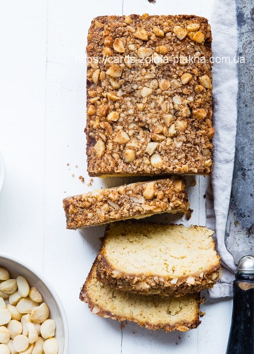 Aufgeschnittener Nuss-Streuselkuchen auf weißem Brett mit Leinentuch, Messer und Schale Macadamianüsse