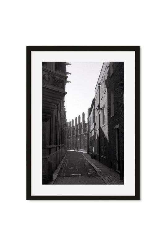 Black-and-white photo of a narrow cobblestone alley between old brick townhouses with chimney stacks